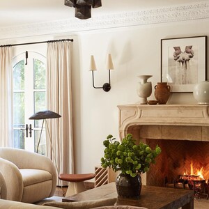 K&ouml;nnte beinhalten: Ein Wohnzimmer-Interieur mit einem Kamin, einem Sofa, einem Couchtisch und einem gro&szlig;en Fenster mit wei&szlig;en Vorh&auml;ngen. Der Kamin hat einen Steinmantel und ein Feuer brennt darin. Das Sofa ist beige und hat ein braunes Kissen. Der Couchtisch ist aus Holz und hat eine Vase mit gr&uuml;nen Blumen darauf. Das Fenster ist gew&ouml;lbt und bietet einen Blick auf einen Garten.