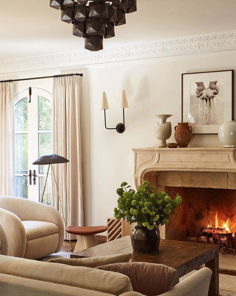 K&ouml;nnte beinhalten: Ein Wohnzimmer-Interieur mit einem Kamin, einem Sofa, einem Couchtisch und einem gro&szlig;en Fenster mit wei&szlig;en Vorh&auml;ngen. Der Kamin hat einen Steinmantel und ein Feuer brennt darin. Das Sofa ist beige und hat ein braunes Kissen. Der Couchtisch ist aus Holz und hat eine Vase mit gr&uuml;nen Blumen darauf. Das Fenster ist gew&ouml;lbt und bietet einen Blick auf einen Garten.