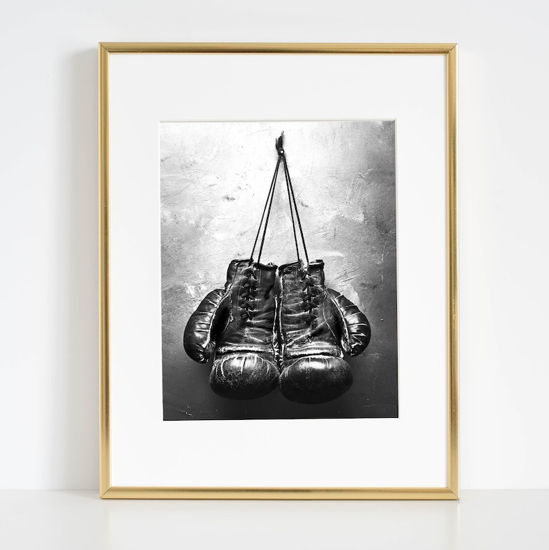 May include: Black and white photo of a pair of leather boxing gloves hanging on a hook against a textured wall.