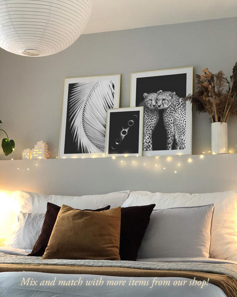 May include: Three black and white framed prints on a wall above a bed with white bedding and brown and black pillows. The prints feature a palm leaf, a celestial image, and two cheetahs.