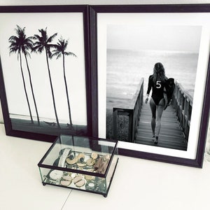 May include: Two black framed prints, one with a black and white photo of palm trees and the other with a black and white photo of a woman walking on a wooden boardwalk towards the ocean. A glass display box with gold and silver jewelry sits on a white surface in front of the prints.