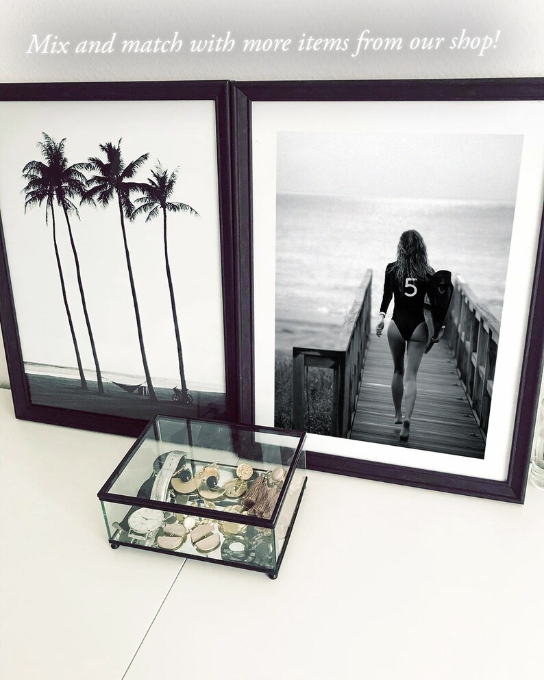 May include: Two black framed prints, one with a black and white photo of palm trees and the other with a black and white photo of a woman walking on a wooden boardwalk towards the ocean. A glass display box with gold and silver jewelry sits on a white surface in front of the prints.
