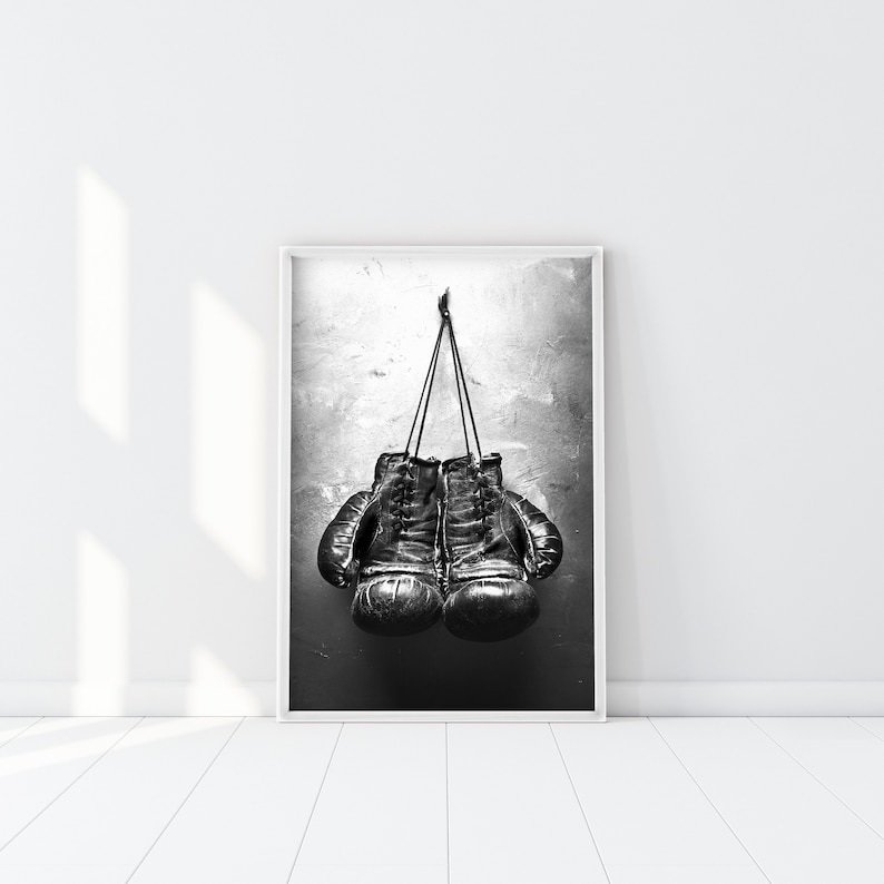 May include: Black and white photograph of a pair of leather boxing gloves hanging on a wall.