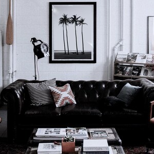 May include: A black leather sofa with three pillows, a black and white patterned pillow, a red and white patterned pillow, and a blue pillow. The sofa is in front of a white brick wall with a framed black and white photo of palm trees on the beach. A coffee table with books and magazines is in front of the sofa.