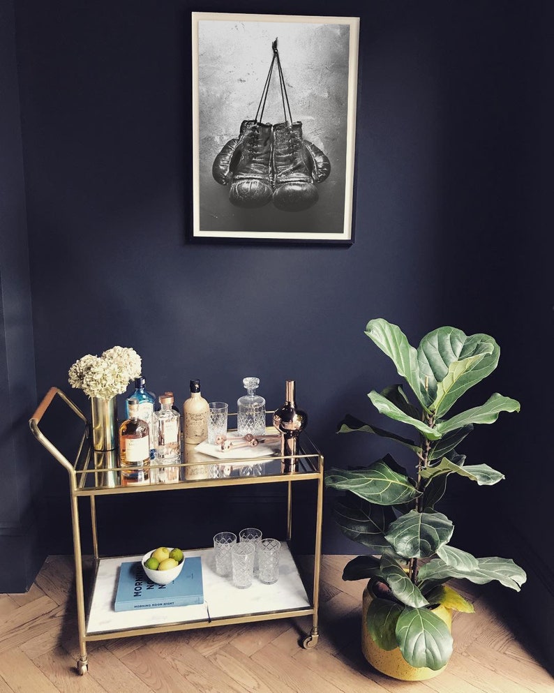 May include: A gold bar cart with a glass top and a marble shelf. The cart is filled with bottles of liquor, glasses, and a plant. A black and white photo of boxing gloves hangs on the wall behind the cart.