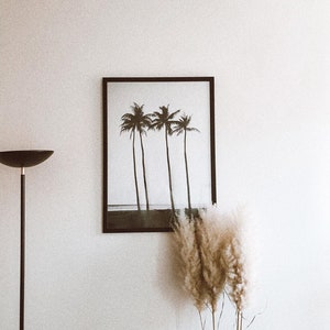May include: A black framed print of four palm trees on a beach with a white background. The print is hanging on a white wall with a black floor lamp and a bouquet of dried pampas grass in the foreground.