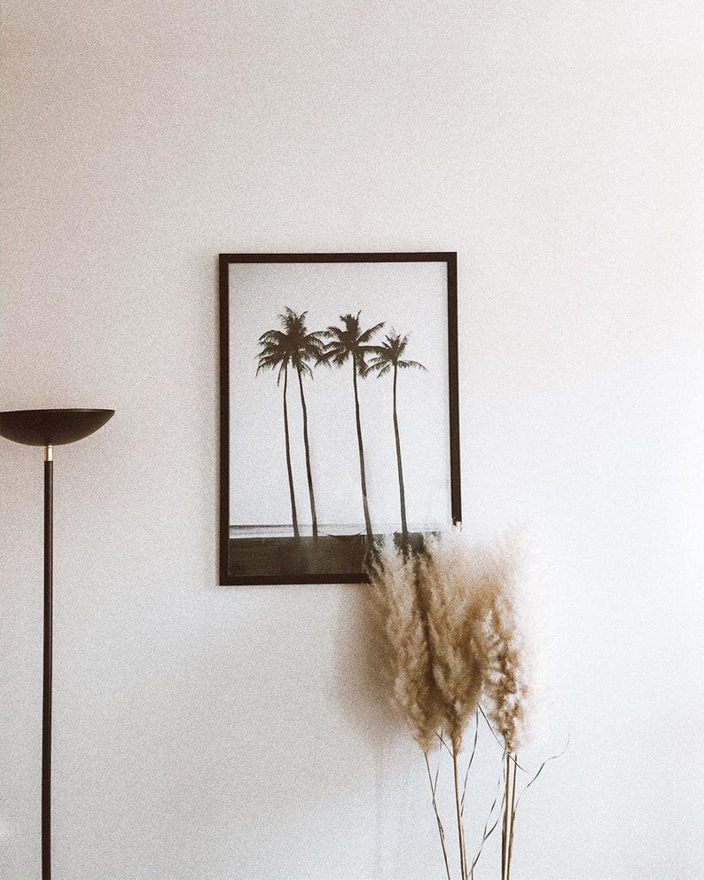May include: A black framed print of four palm trees on a beach with a white background. The print is hanging on a white wall with a black floor lamp and a bouquet of dried pampas grass in the foreground.