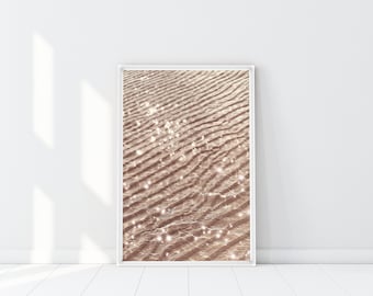 May include: A framed art print featuring a close-up of rippled sand with sparkling highlights. The sand is a warm, light brown color, and the frame is white. The artwork is displayed against a white wall and floor, with natural light creating shadows.