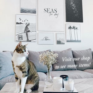 May include: A living room interior with a white couch, a marble coffee table, and a cat sitting on the table. The wall behind the couch is decorated with black and white framed prints of seascapes, palm trees, and waves. The text "SEAS the day" is written on one of the prints. The coffee table has a vase with white flowers, two candles, and a book with the title "CHANEL" on it.