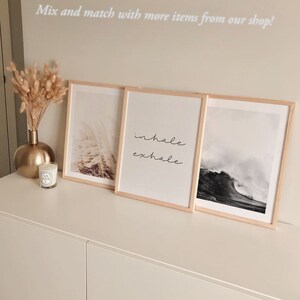 May include: Three framed prints on a white shelf. The left print features a photograph of a field of tall grass. The middle print features the words "inhale exhale" in cursive. The right print features a photograph of a large wave crashing in the ocean.