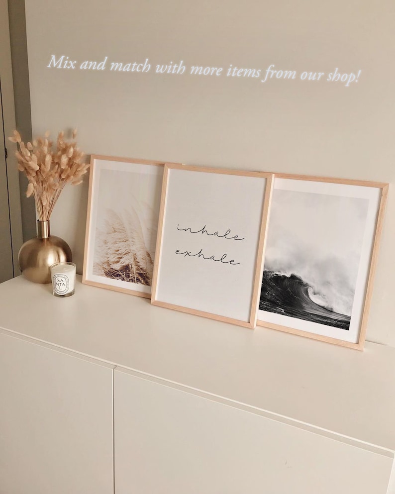 May include: Three framed prints on a white shelf. The left print features a photograph of a field of tall grass. The middle print features the words "inhale exhale" in cursive. The right print features a photograph of a large wave crashing in the ocean.