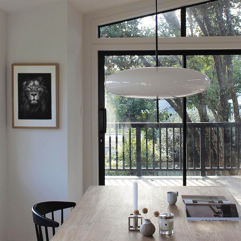 May include: A white oval-shaped pendant light fixture hangs over a wooden dining table with a black chair. The table is decorated with a candle, a vase with dried flowers, and a book. A framed black and white photograph of a lion's face hangs on the wall.