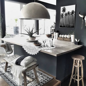 Puede incluir: Una isla de cocina moderna con una encimera blanca y un banco de madera con una manta blanca. Una gran l&aacute;mpara de c&uacute;pula plateada cuelga sobre la isla. Una impresi&oacute;n en blanco y negro de palmeras cuelga en la pared.