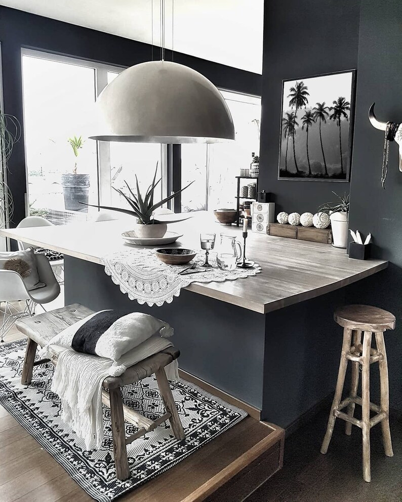 Puede incluir: Una isla de cocina moderna con una encimera blanca y un banco de madera con una manta blanca. Una gran l&aacute;mpara de c&uacute;pula plateada cuelga sobre la isla. Una impresi&oacute;n en blanco y negro de palmeras cuelga en la pared.