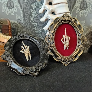 May include: Two ornate frames with black and gold accents, each containing a skeleton hand making the middle finger gesture. One frame has a red velvet background, the other has a black velvet background.