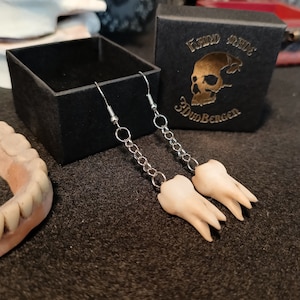 Può includere: Un paio di orecchini d'argento con un ciondolo a forma di dente appeso a ciascuno. Gli orecchini sono in una scatola nera con il testo "Hand Made 3D Bergen" sul coperchio.
