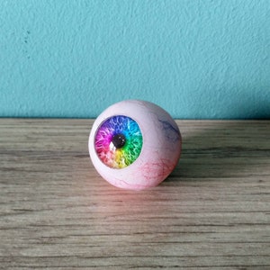 Può includere: Una sfera bianca con un design a occhio arcobaleno. La sfera è testurizzata e ha una leggera sfumatura rosa.