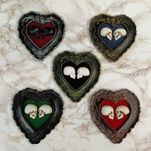 Op de afbeelding: Vijf hartvormige frames met fluwelen interieurs, elk met twee kleine schedels die elkaar aankijken. De frames zijn in verschillende kleuren: zilver, goud, groen, zwart en koper.