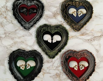Puede incluir: Cinco marcos en forma de corazón con interiores de terciopelo, cada uno con dos cráneos pequeños que se miran. Los marcos son de diferentes colores: plata, oro, verde, negro y cobre.