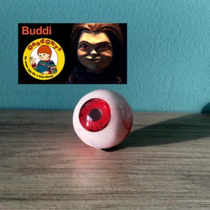 May include: A red and white eyeball prop with a black pupil. The eyeball is sitting on a wooden surface. The text "Buddi" and a logo for "Good Guys" are visible in the background.