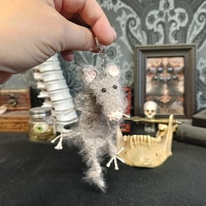 May include: A grey, fluffy rat keyring with pink ears and a pink nose. The rat has small black eyes and white claws. The keyring is held up by a silver ring. The background includes a framed picture, a skull, and a jawbone.