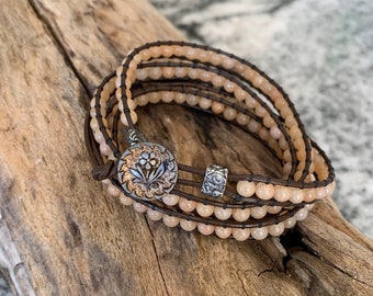May include: A multi-strand bracelet with light brown beads and brown leather cord. The bracelet has a silver and gold floral button closure.