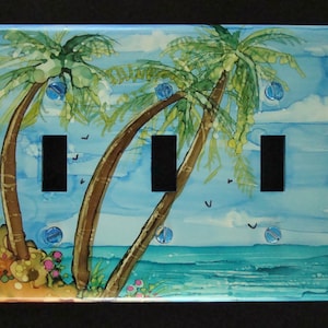 May include: A light blue, hand-painted light switch cover with a tropical scene. The cover features three palm trees, a blue sky with white clouds, and a turquoise ocean. The switch cover is designed for three light switches.