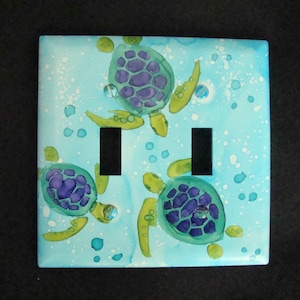 May include: A light blue double light switch cover with a painted design of green and purple sea turtles swimming in a blue ocean with white bubbles.