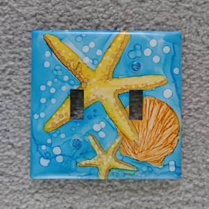 May include: A light blue double light switch cover with a painted design of two yellow starfish and a seashell on a blue background with white bubbles.