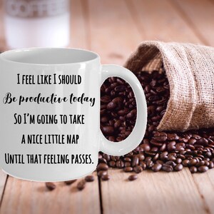 Puede incluir: Taza de caf&eacute; de cer&aacute;mica blanca con una cita humor&iacute;stica en texto negro: "I FEEL LIKE I SHOULD Be productive today SO I'M GOING TO TAKE A NICE LITTLE NAP UNTIL THAT FEELING PASSES." Granos de caf&eacute; y un saco de yute en el fondo.