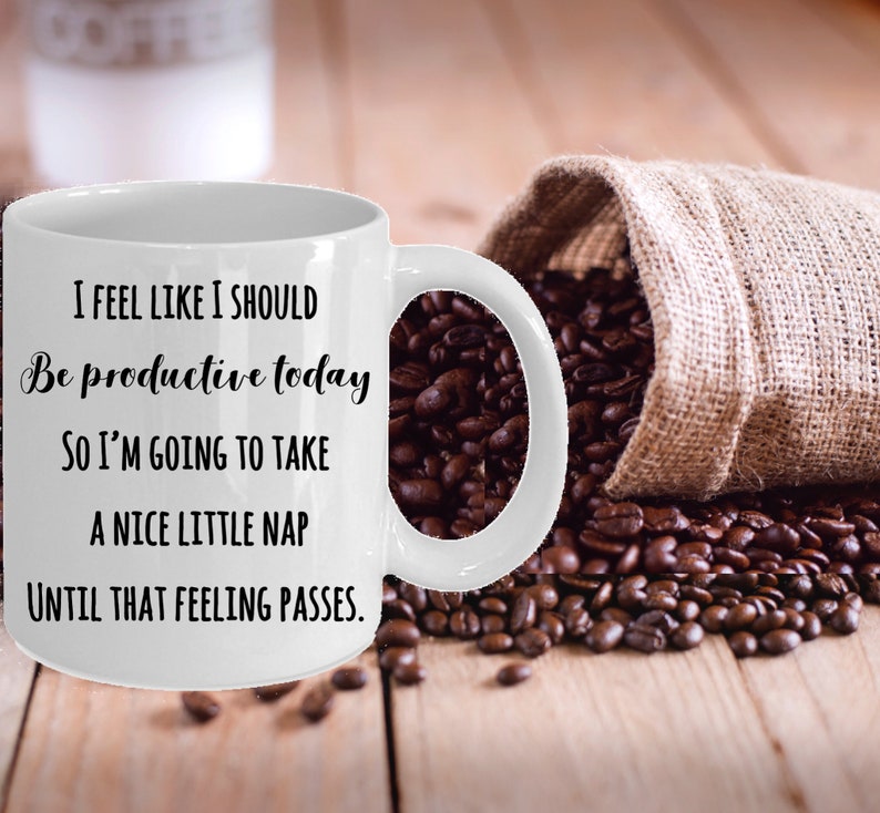 Puede incluir: Taza de caf&eacute; de cer&aacute;mica blanca con una cita humor&iacute;stica en texto negro: "I FEEL LIKE I SHOULD Be productive today SO I'M GOING TO TAKE A NICE LITTLE NAP UNTIL THAT FEELING PASSES." Granos de caf&eacute; y un saco de yute en el fondo.
