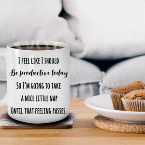 Puede incluir: Taza de cer&aacute;mica blanca llena de caf&eacute;, con el texto "I FEEL LIKE I SHOULD Be productive today SO I'M GOING TO TAKE A NICE LITTLE NAP UNTIL THAT FEELING PASSES." La taza est&aacute; sobre un posavasos, con magdalenas en un plato.