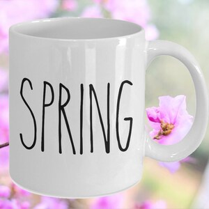 Puede incluir: Taza de cer&aacute;mica blanca con la palabra "SPRING" en negro, con letras manuscritas. La taza tiene un asa curva y se encuentra sobre un fondo borroso de flores rosas y follaje verde.