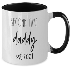 May include: White ceramic mug with a black interior and handle. The mug has the words "SECOND TIME daddy est. 2021" printed in black script. A gift for a new father.