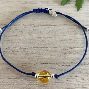 Könnte beinhalten: Handgefertigtes Armband mit einer leuchtend gelben Edelsteinperle, silberfarbenen Metallperlen und einem Federanhänger. Das marineblaue Band hat verstellbare Knoten für eine bequeme Passform.