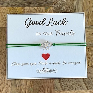 Könnte beinhalten: Ein grünes Armband aus Schnur mit einem silbernen Glückskleeblatt-Anhänger. Das Armband befindet sich auf einer weißen Karte mit dem Text "Good Luck on Your Travels" und "Close your eyes. Make a wish. Be amazed." Die Karte hat auch ein rotes Herz und das Logo "destinii HANDMADE JEWELLERY".