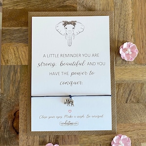 Puede incluir: Una tarjeta con una ilustración de elefante y el texto "A LITTLE REMINDER YOU ARE strong, beautiful AND YOU HAVE THE power to conquer." Una pulsera de cuerda azul con un dije de elefante debajo. Adornos florales rosas sobre madera.