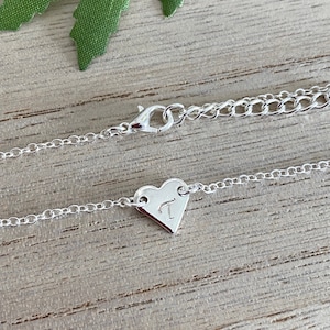 Peut inclure: Un collier en argent avec un pendentif en forme de cœur. Le pendentif porte la lettre "T" gravée. Le collier est posé sur une surface en bois. La chaîne est délicate et possède un fermoir homard.