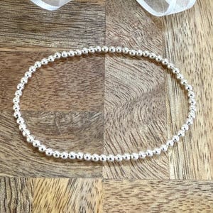 May include: A silver beaded bracelet with a simple design. The bracelet is made of small, round beads that are strung together on a thin wire. The bracelet is lying flat on a wooden surface.