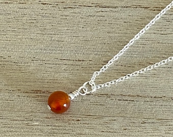 May include: A silver necklace featuring a small, round, orange-brown gemstone pendant. The pendant is wire-wrapped and suspended from a delicate silver chain. The necklace is displayed on a wooden surface.
