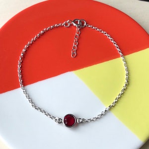 May include: A delicate silver bracelet featuring a single, round, deep red gemstone. The bracelet has a fine chain design and an adjustable clasp. The jewelry is displayed on a plate with red, yellow, and white sections.