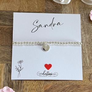 Könnte beinhalten: Ein silbernes Perlenarmband mit einem kleinen runden Anhänger. Der Anhänger hat einen Buchstaben. Das Armband wird auf einer weißen Karte mit dem Namen "Sandra" in Schreibschrift präsentiert. Ein rotes Herz und der Markenname "destinii" sind ebenfalls auf der Karte.