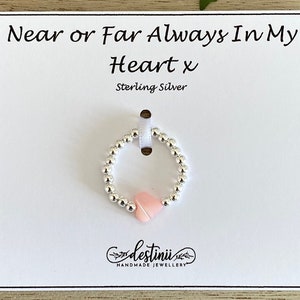 May include: A silver beaded bracelet with a pink heart charm. The bracelet is displayed on a white card with the text "Near or Far Always In My Heart x Sterling Silver" and the logo "destinni HANDMADE JEWELLERY".