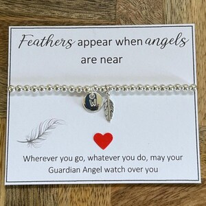 Op de afbeelding: Een zilveren kralenarmband met een bedel met de letter "W" en een veertje. De kaart achter de armband heeft de tekst "Feathers appear when angels are near" en "Wherever you go, whatever you do, may your Guardian Angel watch over you."