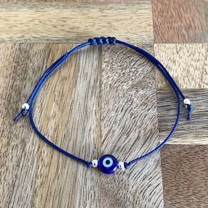 Puede incluir: Un brazalete de ojo turco azul y blanco con un cordón azul oscuro. El brazalete es ajustable y tiene cuentas plateadas a cada lado del amuleto.