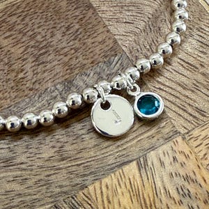May include: Silver beaded bracelet featuring a charm with the letter "J" and a teal gemstone. The bracelet is presented on a wooden surface.
