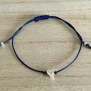 Puede incluir: Un brazalete de cuerda azul marino con un dije de corazón plateado y detalles de cuentas plateadas.
