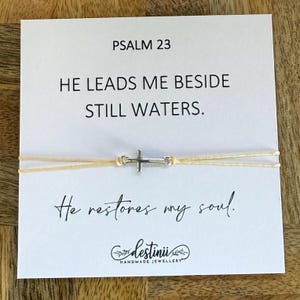 Könnte beinhalten: Ein cremefarbenes Kordelarmband mit einem silbernen Kreuzanhänger, präsentiert auf einer weißen Karte. Die Karte zeigt den Text "PSALM 23" und das Zitat "Er führt mich zu stillen Wassern. Er erquickt meine Seele."