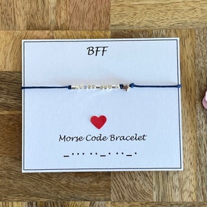 Puede incluir: Una tarjeta blanca con la palabra "BFF" en texto negro. Un brazalete de cuerda azul con cuentas plateadas y un dije de estrella está en la tarjeta. La tarjeta también dice "Pulsera de código Morse" y muestra el código para BFF en código Morse.