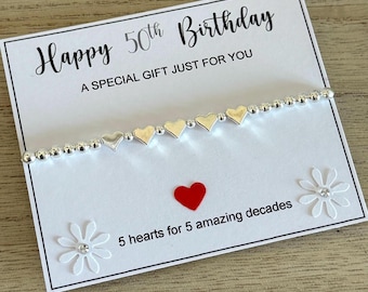 May include: A white card with the text "Happy 50th Birthday" and "A special gift just for you". A silver bracelet with five silver heart charms is on the card. The card also has a red heart and two white flowers with rhinestones. The text "5 hearts for 5 amazing decades" is on the card.